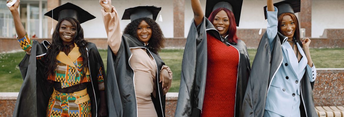 Excited african american female students at their graduation. Group of young afro american female student dressed in black graduation gown. Campus as a background. Girls cheerfully smiling with arms up, holding diploma.